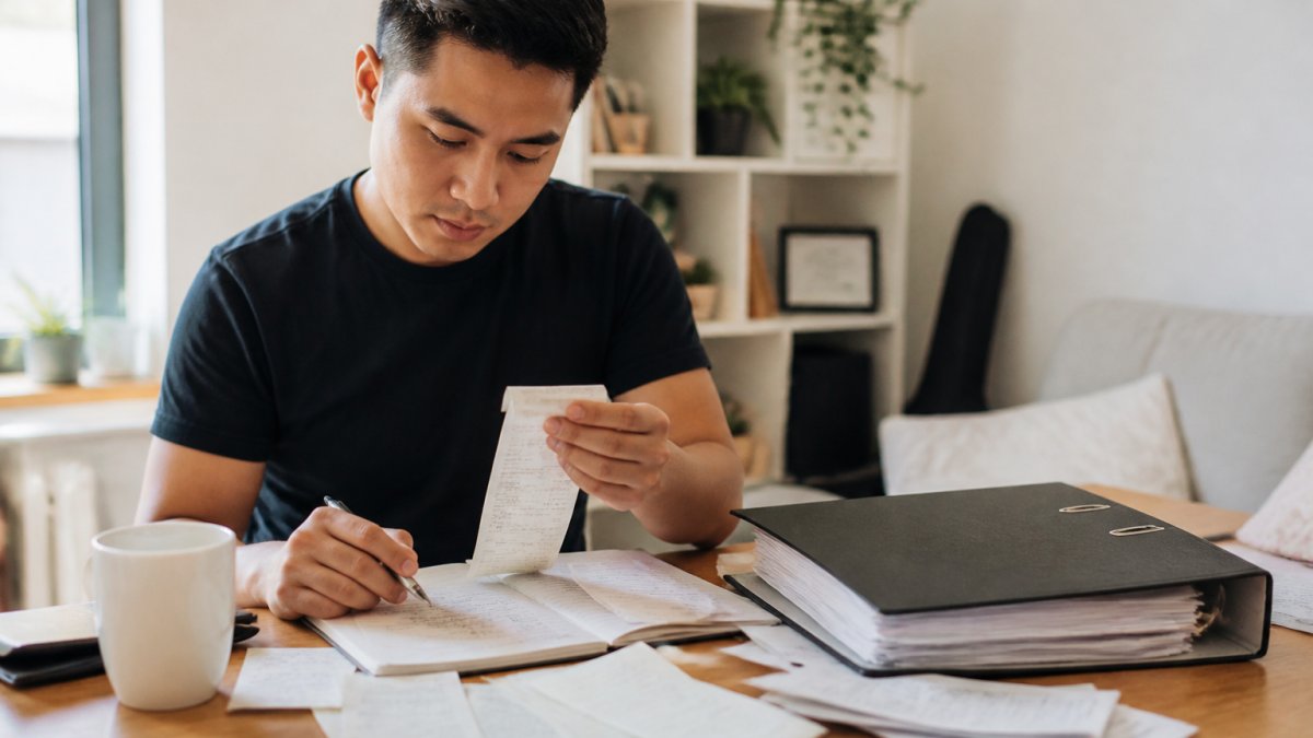 A startup founder at a desk sorting receipts and reviewing expense categories on a laptop, with a folder of organized documents and a coffee mug nearby in a bright home office